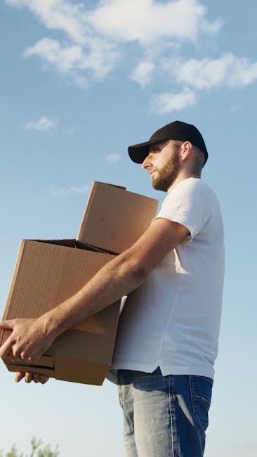 A man wearing a black cap, white t-shirt, and jeans is standing outdoors against a backdrop of a blue sky with scattered clouds, during daylight hours. He is holding two cardboard boxes, one larger and one smaller, which appear to contain household items. The larger box is being supported with both arms, while he grasps the smaller box on top with one hand. The scene suggests a home relocation or packing process, with the man preparing to load or unload the boxes. Visible in the background are the exterior of a property, possibly with entry steps or a pavement area, indicating an indoor-to-outdoor furniture transport or moving activity. This image is associated with [COMPANY_NAME] and their removal and moving services, including packing and loading, as part of house relocation in Colliers Wood.