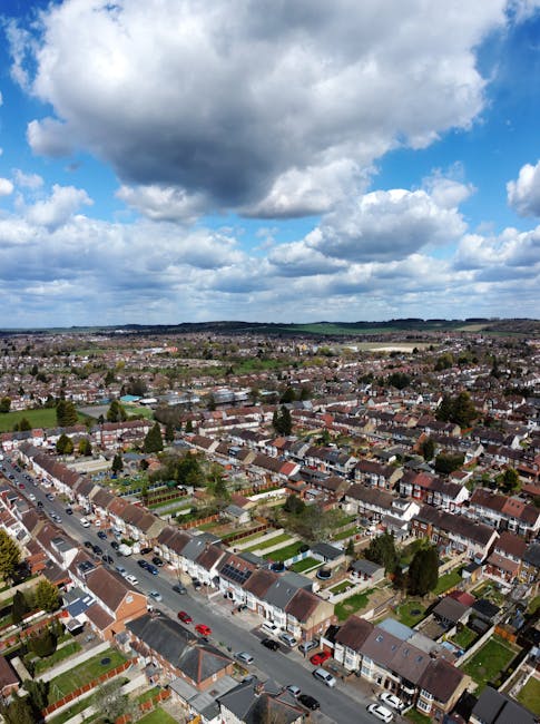 An aerial view of a suburban residential area with densely packed houses, terraced homes, and small gardens, situated along a wide street with parked cars. The scene is captured during daytime under a partly cloudy sky with large, dark clouds overhead. In the foreground, there are houses with pitched roofs, some with visible driveways and parked vehicles. The middle ground shows more houses extending into the distance, with a mix of greenery and built-up areas. The background features rolling hills and a horizon under a sky filled with dramatic cloud formations. This comprehensive view illustrates a typical UK neighbourhood, suitable for visual context related to house removals, furniture transport, and home relocation services provided by Man and Van Colliers Wood during packing and loading processes.